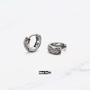 May include: A pair of silver-toned hoop earrings featuring an embossed dragon scale pattern. The earrings have a hinged closure mechanism and a polished metallic finish. The text Black Fox with a small fox icon is visible below the earrings.