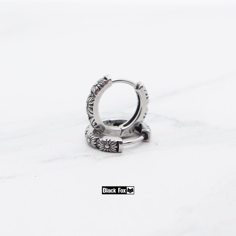 May include: Silver hoop earrings with a floral pattern. The earrings are made of a shiny metal and have a detailed design of small flowers around the hoops. The brand name "Black Fox" is in a black rectangle.