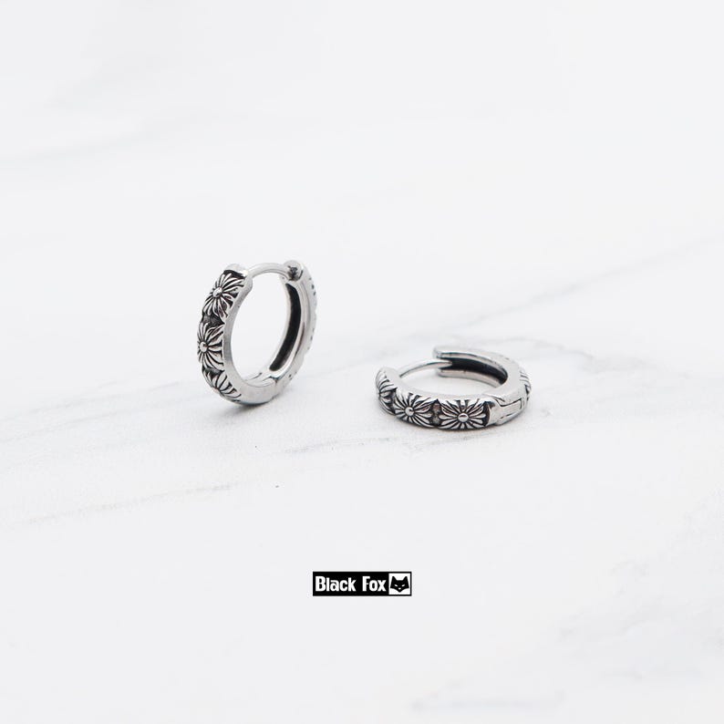 May include: A pair of silver hoop earrings featuring a floral pattern. The earrings are small, circular, and adorned with flower-shaped engravings. They are presented on a white marble surface. The brand name "Black Fox" is visible.