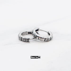May include: A pair of silver hoop earrings with a floral pattern. The earrings have a shiny finish and are decorated with a repeating design of small flowers. The background is white. The brand name "Black Fox" is visible.