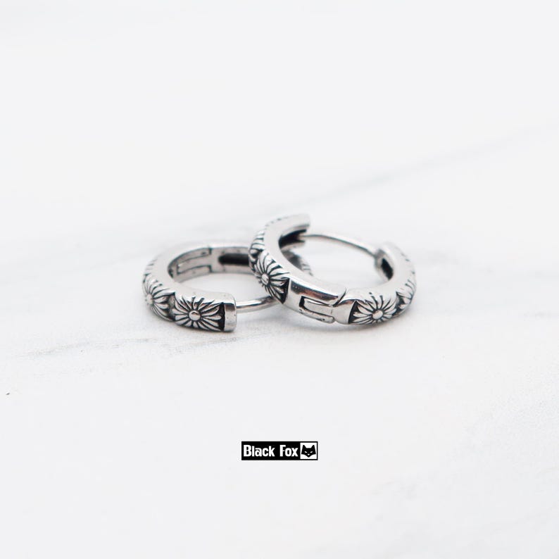 May include: A pair of silver hoop earrings with a floral pattern. The earrings have a shiny finish and are decorated with a repeating design of small flowers. The background is white. The brand name "Black Fox" is visible.