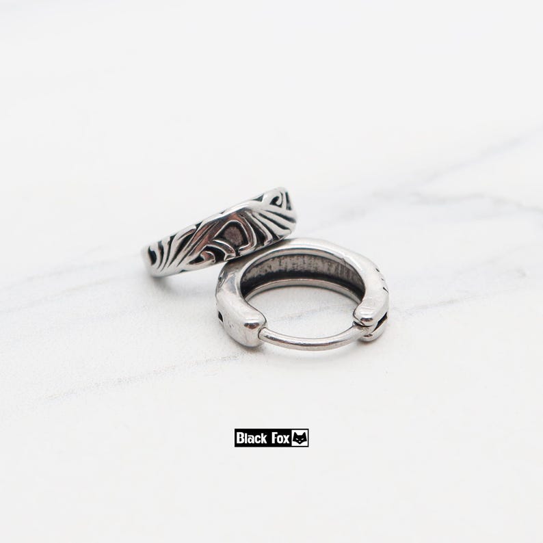 May include: Two silver-colored hoop earrings with an engraved design. One earring is open, displaying the clasp, while the other is closed. The earrings are on a white marble surface. The text "Black Fox" is at the bottom.