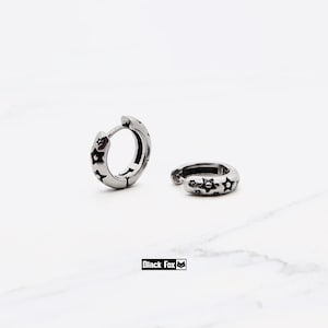 May include: A pair of silver-toned hoop earrings featuring a repeating pattern of black-filled stars around the outer surface. One earring stands upright while the other lies flat. The text Black Fox is visible at the bottom of the frame.