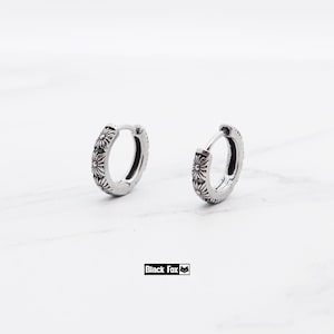May include: Two silver hoop earrings with a floral pattern. The earrings are small and have a hinged clasp. The earrings are set against a white backdrop. The brand name "Black Fox" is printed in a black rectangle.