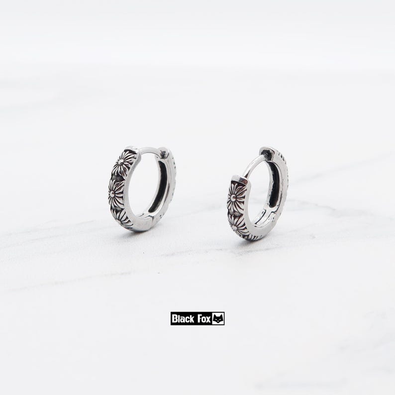 May include: Two silver hoop earrings with a floral pattern. The earrings are small and have a hinged clasp. The earrings are set against a white backdrop. The brand name "Black Fox" is printed in a black rectangle.