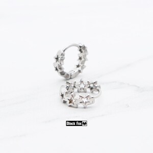 May include: Two silver hoop earrings with star-shaped cutouts. The earrings are displayed on a white surface. The text "Black Fox" is in a black rectangle at the bottom of the image.