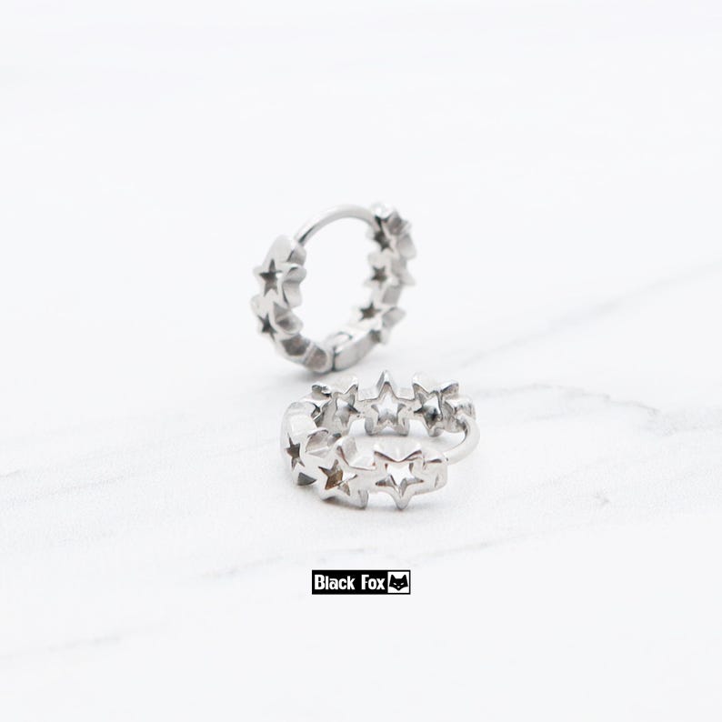 May include: Two silver hoop earrings with star-shaped cutouts. The earrings are displayed on a white surface. The text "Black Fox" is in a black rectangle at the bottom of the image.
