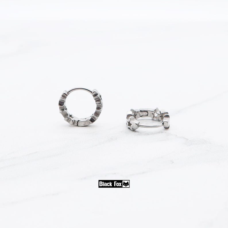 May include: Two silver hoop earrings with star-shaped cutouts. The earrings are displayed on a white marble surface. The brand name "Black Fox" is printed in a black rectangle.