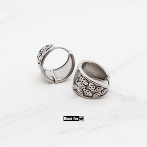 May include: Two silver-colored hoop earrings. Each earring features a repeating skull pattern around the outer edge. The earrings are displayed on a white marble surface. The brand name "Black Fox" is visible.