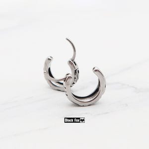 May include: A pair of silver-colored hoop earrings with intricate detailing. The earrings are open, showcasing their design and texture. The brand name "Black Fox" is displayed in a small black rectangle.