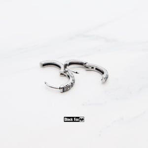 May include: Silver-coloured hoop earrings with a floral pattern. The earrings have a hinged closure and are displayed on a white marble surface. The text "Black Fox" is visible. The earrings are a stylish accessory.