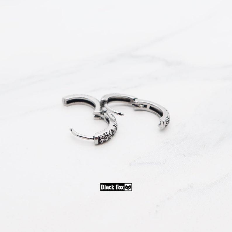 May include: Silver-coloured hoop earrings with a floral pattern. The earrings have a hinged closure and are displayed on a white marble surface. The text "Black Fox" is visible. The earrings are a stylish accessory.