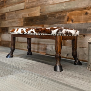 May include: A rustic bench with a cowhide seat and dark wooden legs shaped like horse hooves. Two vintage metal milk cans stand nearby, adding to the Western theme. The bench is approximately 36 inches long.