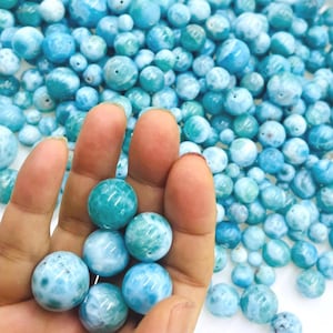 以下が含まれることがあります： さまざまなサイズの丸い青緑色の宝石ビーズのコレクション。 いくつかのビーズは手で持たれ、他のビーズは白いトレイを満たしています。 ビーズはマーブル模様で、明るい色と暗い色の青色の渦があります。