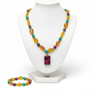 Puede incluir: Un conjunto de collar y pulsera con cuentas ovaladas de color ámbar, intercaladas con cuentas turquesas y moradas. El collar tiene un colgante rectangular granate. Las joyas se exhiben sobre un busto blanco.