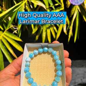 K&ouml;nnte beinhalten: Ein hellblaues Larimar-Armband mit runden Perlen wird in einer kleinen Schachtel pr&auml;sentiert. Der Text "High Quality AAA Larimar Bracelet" ist sichtbar. Das Armband wird in der Hand einer Person gehalten, mit einem Hintergrund aus gr&uuml;nen Bl&auml;ttern.