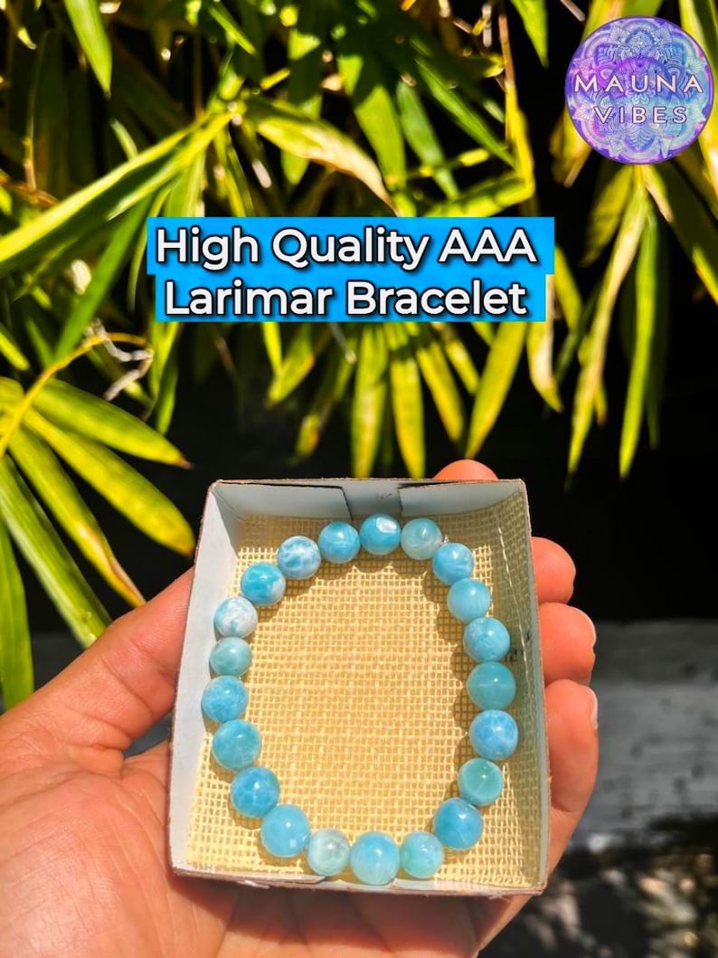 K&ouml;nnte beinhalten: Ein hellblaues Larimar-Armband mit runden Perlen wird in einer kleinen Schachtel pr&auml;sentiert. Der Text "High Quality AAA Larimar Bracelet" ist sichtbar. Das Armband wird in der Hand einer Person gehalten, mit einem Hintergrund aus gr&uuml;nen Bl&auml;ttern.