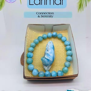 K&ouml;nnte beinhalten: Ein Larimar-Armband und ein gro&szlig;er Larimar-Stein in einer kleinen Schachtel. Das Armband besteht aus runden, hellblauen Perlen. Die Schachtel hat eine gewebte Textur und den Text "Larimar, Connection & Serenity".