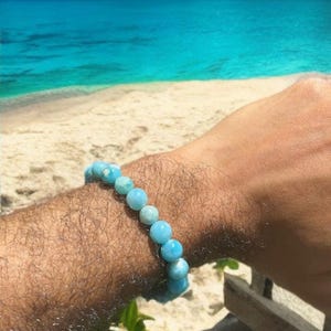 K&ouml;nnte beinhalten: Ein hellblaues Perlenarmband wird am Handgelenk getragen. Das Armband besteht aus runden, hellblauen Perlen. Der Hintergrund zeigt einen Strand, das Meer und den Himmel. Ein kreisf&ouml;rmiges Logo mit dem Text "MAUNA VIBES" befindet sich in der oberen rechten Ecke.
