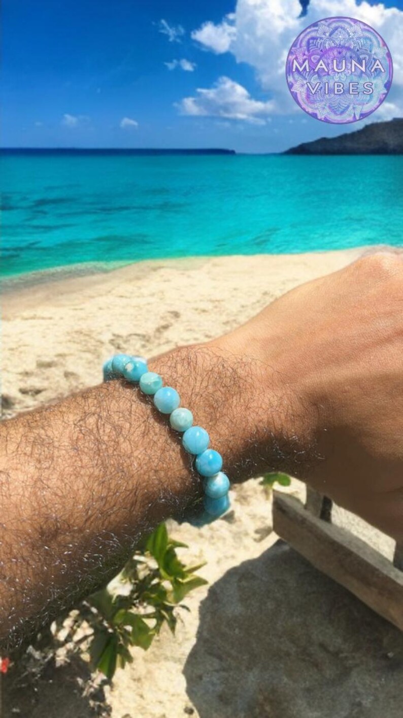 K&ouml;nnte beinhalten: Ein hellblaues Perlenarmband wird am Handgelenk getragen. Das Armband besteht aus runden, hellblauen Perlen. Der Hintergrund zeigt einen Strand, das Meer und den Himmel. Ein kreisf&ouml;rmiges Logo mit dem Text "MAUNA VIBES" befindet sich in der oberen rechten Ecke.