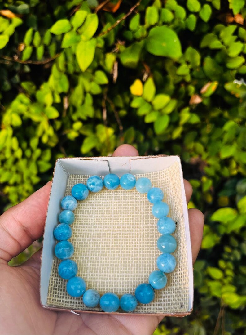 K&ouml;nnte beinhalten: Ein hellblaues Perlenarmband, pr&auml;sentiert in einer kleinen, offenen Schachtel mit beigem Stofffutter. Das Armband besteht aus runden, polierten Perlen. Der Hintergrund ist eine verschwommene Ansicht von gr&uuml;nem Laub.