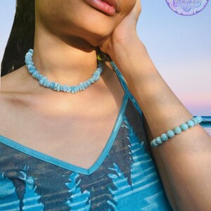 K&ouml;nnte beinhalten: Ein hellblaues Perlenkette- und Armband-Set. Die Halskette ist im Choker-Stil gehalten, und das Armband besteht aus einer Reihe runder Perlen. Die Frau tr&auml;gt ein blaues Tanktop mit einem Baumdesign und dem Text "MAUNA VIBES" in einem Kreis.