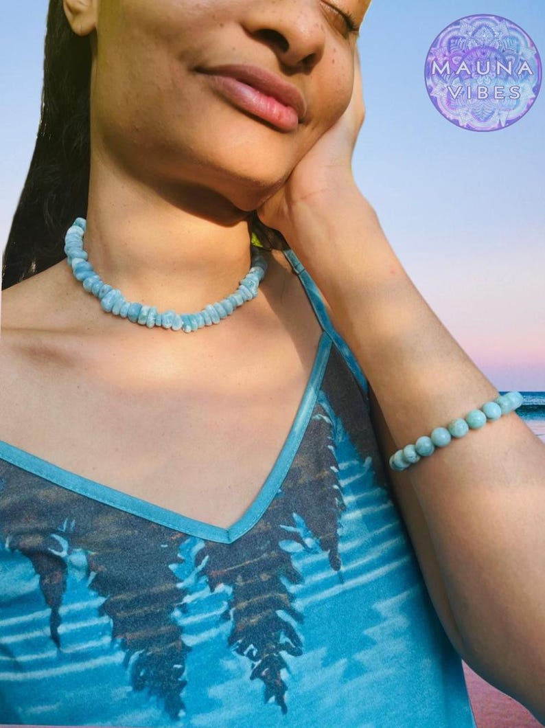 K&ouml;nnte beinhalten: Ein hellblaues Perlenkette- und Armband-Set. Die Halskette ist im Choker-Stil gehalten, und das Armband besteht aus einer Reihe runder Perlen. Die Frau tr&auml;gt ein blaues Tanktop mit einem Baumdesign und dem Text "MAUNA VIBES" in einem Kreis.