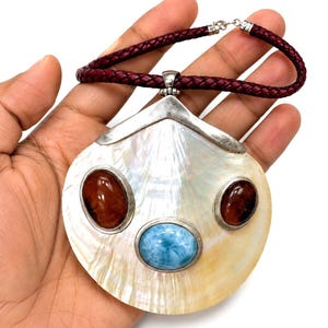 May include: A large, pearlescent pendant necklace with a silver-tone metal setting. The pendant features three oval gemstones: two brown and one light blue. The necklace has a braided, burgundy leather cord.