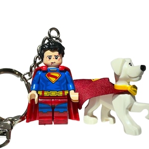 May include: A Superman-themed keyring featuring a Lego Superman figure with a red cape and a white dog with a red cape. The Superman figure has a blue and red suit and a silver keyring.