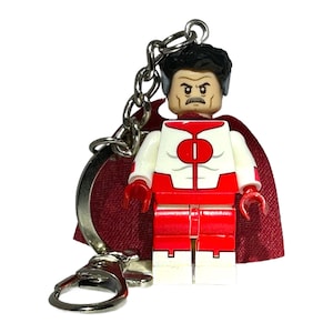 May include: A Lego-style keychain of a superhero figure in a red and white costume, with black hair and a serious expression. The figure has a red cape and is attached to a silver chain and clasp.