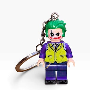May include: A keyring featuring a plastic minifigure of a character with bright green hair, white face paint, dark eye makeup, and a red smile. The figure wears a purple jacket over a lime green waistcoat, a patterned shirt, a tie, and purple pinstriped trousers.
