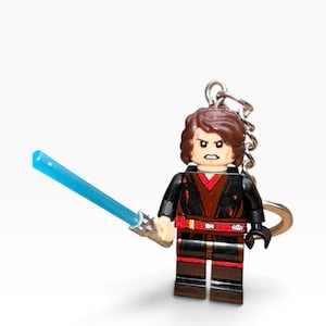 May include: A LEGO minifigure keyring featuring a character with brown hair, a scar over the right eye, and an angry expression. The figure wears a black and dark red tunic with a red belt and holds a translucent blue plastic lightsaber accessory.