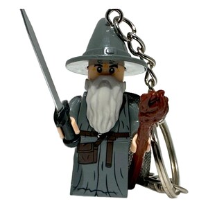 May include: A grey Lego wizard figure with a long white beard, holding a silver sword and a brown staff. The figure wears a grey hat and robe, and is attached to a keyring.