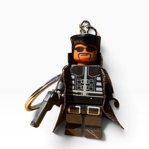 May include: A plastic minifigure keyring featuring a character with a tan complexion, black sunglasses, and a goatee. The figure wears a black trench coat over a black torso with silver detailing and black trousers, holding a black plastic pistol.