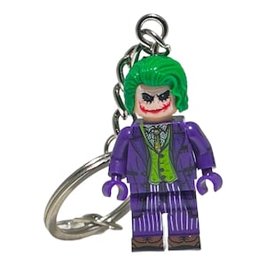 May include: A Joker-themed keyring featuring a Lego-style figure. The figure has green hair, a painted face, and wears a purple suit with a green waistcoat and striped trousers. The keyring includes a silver chain and a key ring.