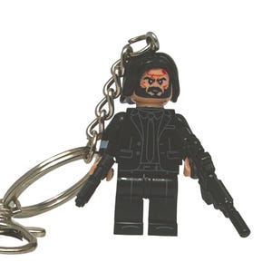 May include: A black Lego-style keyring featuring a minifigure in a black suit, with a beard and a bloodied face. The figure holds two black rifles. The keyring has a silver chain and clasp.