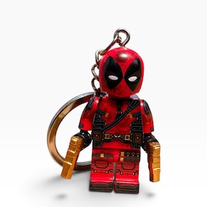 May include: A Deadpool character keyring featuring a red and black suit with detailed tactical gear, a mask with white eye patches, and two gold-coloured toy pistols held in each hand. The figure is attached to a silver metal keyring and chain.