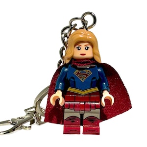 May include: A Supergirl Lego figure keyring. The figure has a blue and red outfit with a red cape and blonde hair. The keyring includes a silver chain and clasp.