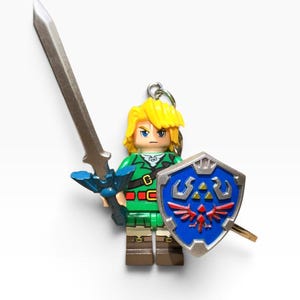 May include: A keyring featuring a plastic minifigure of a character with blonde hair, wearing a green tunic and brown boots. The figure holds a silver sword in one hand and a blue shield with a red bird and gold triangle emblem in the other.
