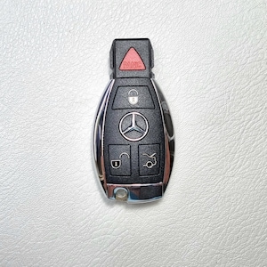 May include: Black and silver car key with a red "PANIC" button. The key has buttons for locking, unlocking, and other functions, with the Mercedes-Benz logo prominently displayed. The key is on a white surface.