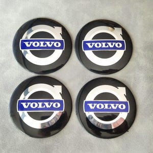 Könnte beinhalten: Vier schwarze Volvo Radnabenabdeckungen. Jede Kappe zeigt das Volvo-Logo in Weiß und Silber, mit dem Wort "VOLVO" in Weiß auf blauem rechteckigem Hintergrund. Die Kappen sind kreisförmig und haben eine glänzende Oberfläche.