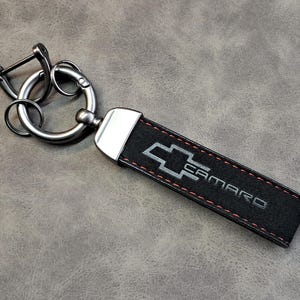Op de afbeelding: Zwarte leren sleutelhanger met een zilveren metalen ring en een zilveren Chevrolet Camaro-logo met rode stiksels.