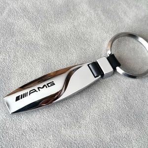 Può includere: Un portachiavi argentato con un cinturino in pelle nera e un anello per chiavi argentato. Il portachiavi ha un'etichetta in metallo argentato con la parola "AMG" in lettere nere.