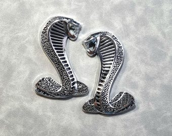 May include: Two silver-colored metal Cobra emblems. Each emblem features a detailed snake design with a raised, textured pattern.