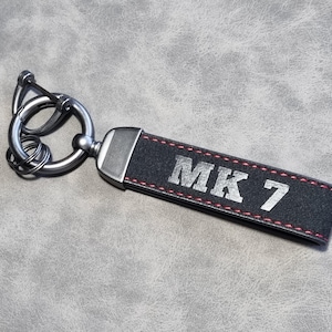 以下が含まれることがあります： 赤いステッチと銀色の金属リングが付いた黒い革のキーホルダー。キーホルダーには銀色のエンボス加工で「MK 7」という文字が刻まれています。