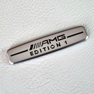 Puede incluir: Una insignia de metal plateado con letras negras que dice "AMG EDITION 1".
