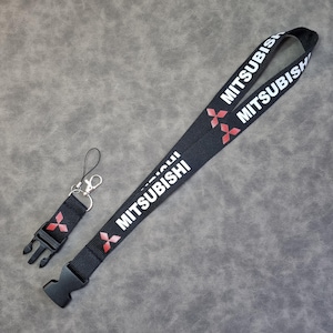 Peut inclure: Lanière noire avec du texte blanc qui dit "MITSUBISHI" et un logo Mitsubishi rouge. Un porte-clés noir avec un clip argenté est attaché à la lanière.