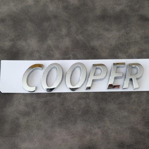 May include: Shiny chrome "COOPER" emblem, likely for a car, mounted on a white strip against a textured gray background. The letters are bold and reflect light, suggesting a polished finish.