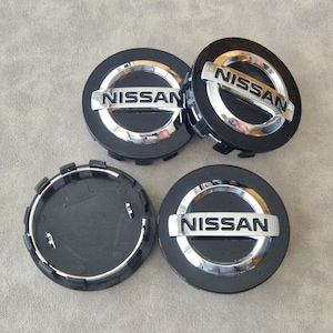 Puede incluir: Cuatro tapacubos Nissan negros y plateados. Cada tapacubo presenta la palabra "NISSAN" en letras plateadas sobre un fondo negro. Los tapacubos son redondos con un borde plateado.
