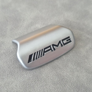 Può includere: Emblema AMG argentato con scritte nere e una serie di linee angolate. L'emblema ha una forma curva ed è posto su uno sfondo grigio strutturato. Il logo AMG è ben visibile.